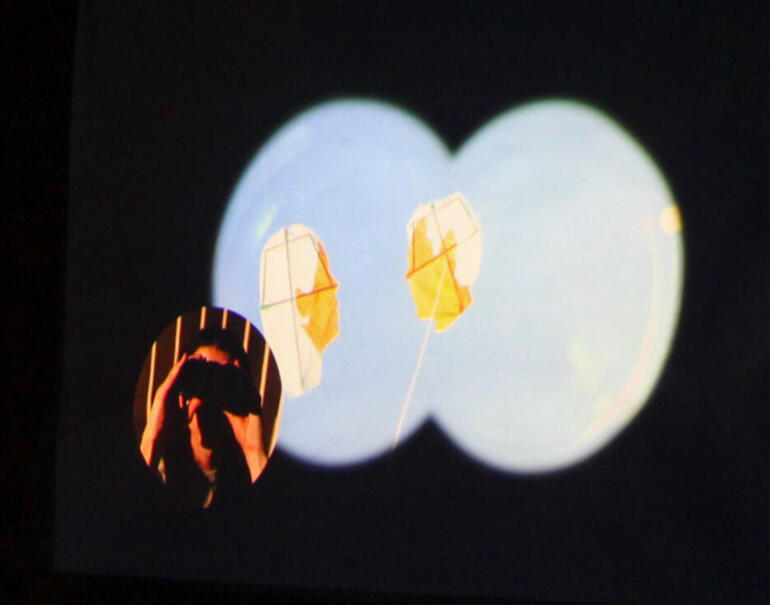 Still from video by Lane Sheehy. Features binocular view for two kites floating in the sky with a smaller circle to the left of the artist holding binoculars to her eyes. Piece is photographed at night and projected onto a temporary wall.