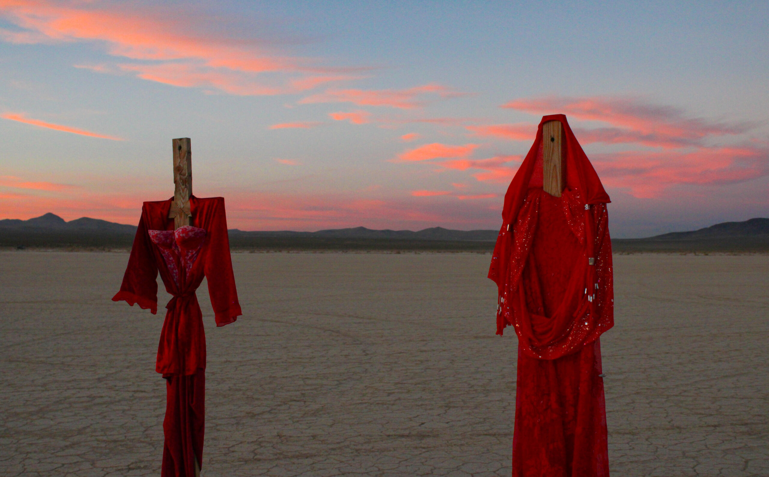 Image of sculpture by Nishan Ganimian and Romina Villareal. Features two tall sculptures with sunset in background, sculptures are wooden poles with red dressage adorning them, resembling figures.