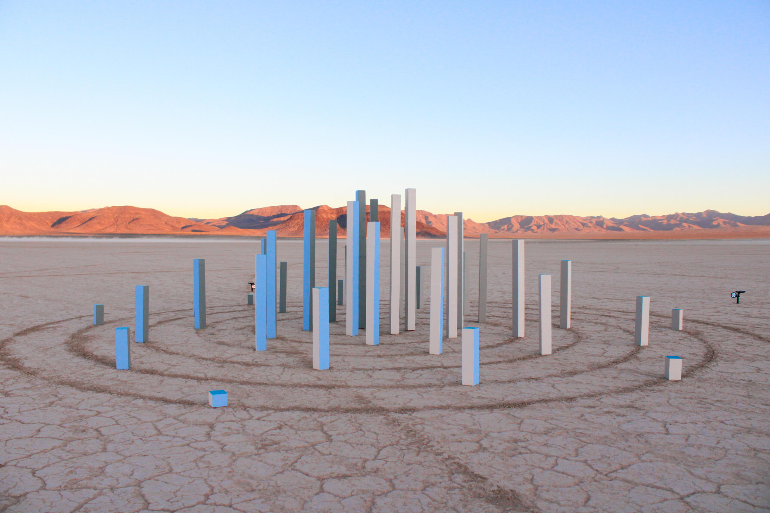 Image of sculpture by Abney Wallace. Features vertical pieces of dimensional lumber painted various shades of white and blue arranged in a crop circle with circular lines scratched into lake-bed.