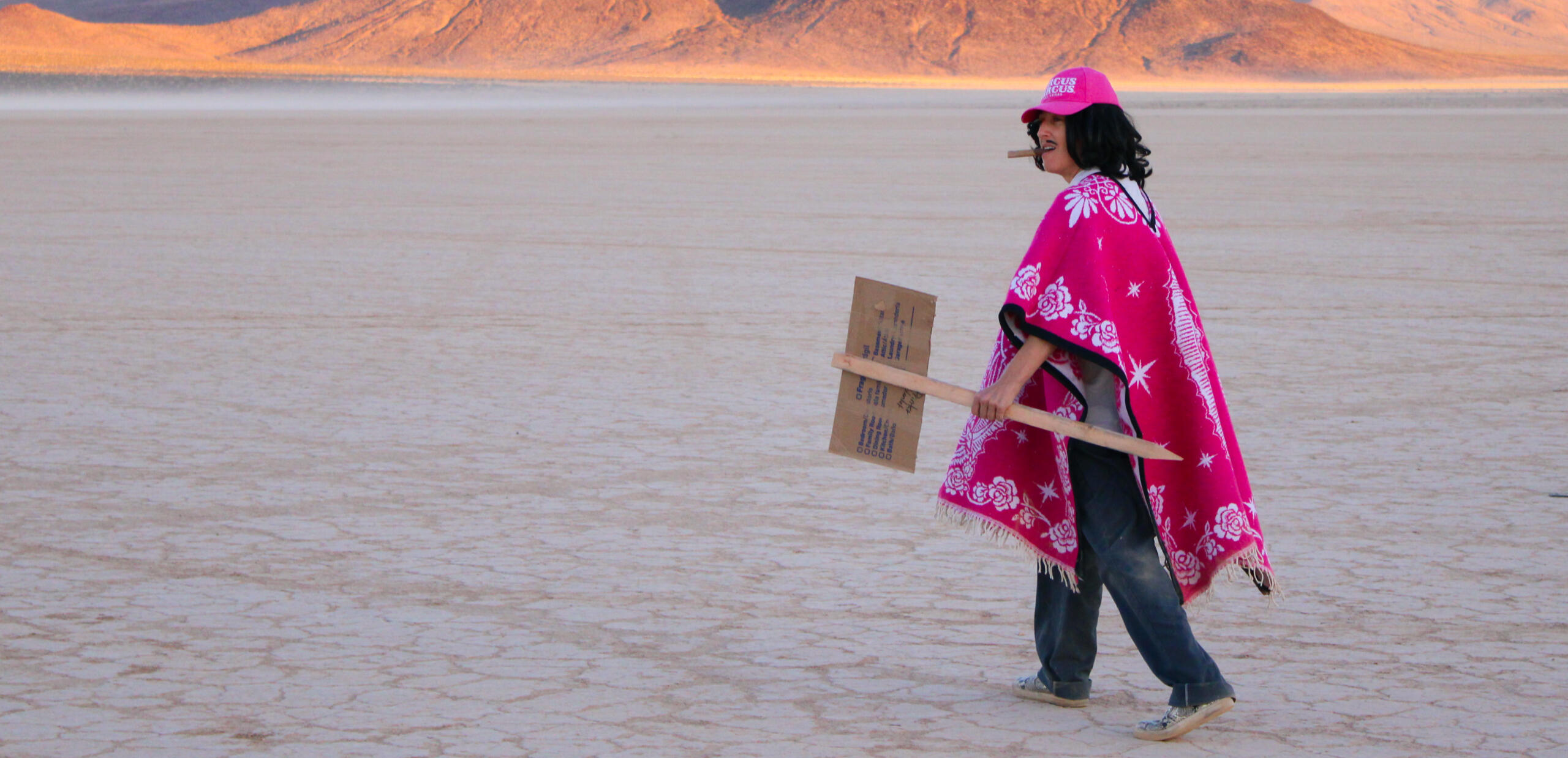 Still from durational performance by Adriana Chavez. Features Adriana dressed as her character Juan Chico with cigar in mouth, walking with cardboard sign that says "claimed" on it in black letters.