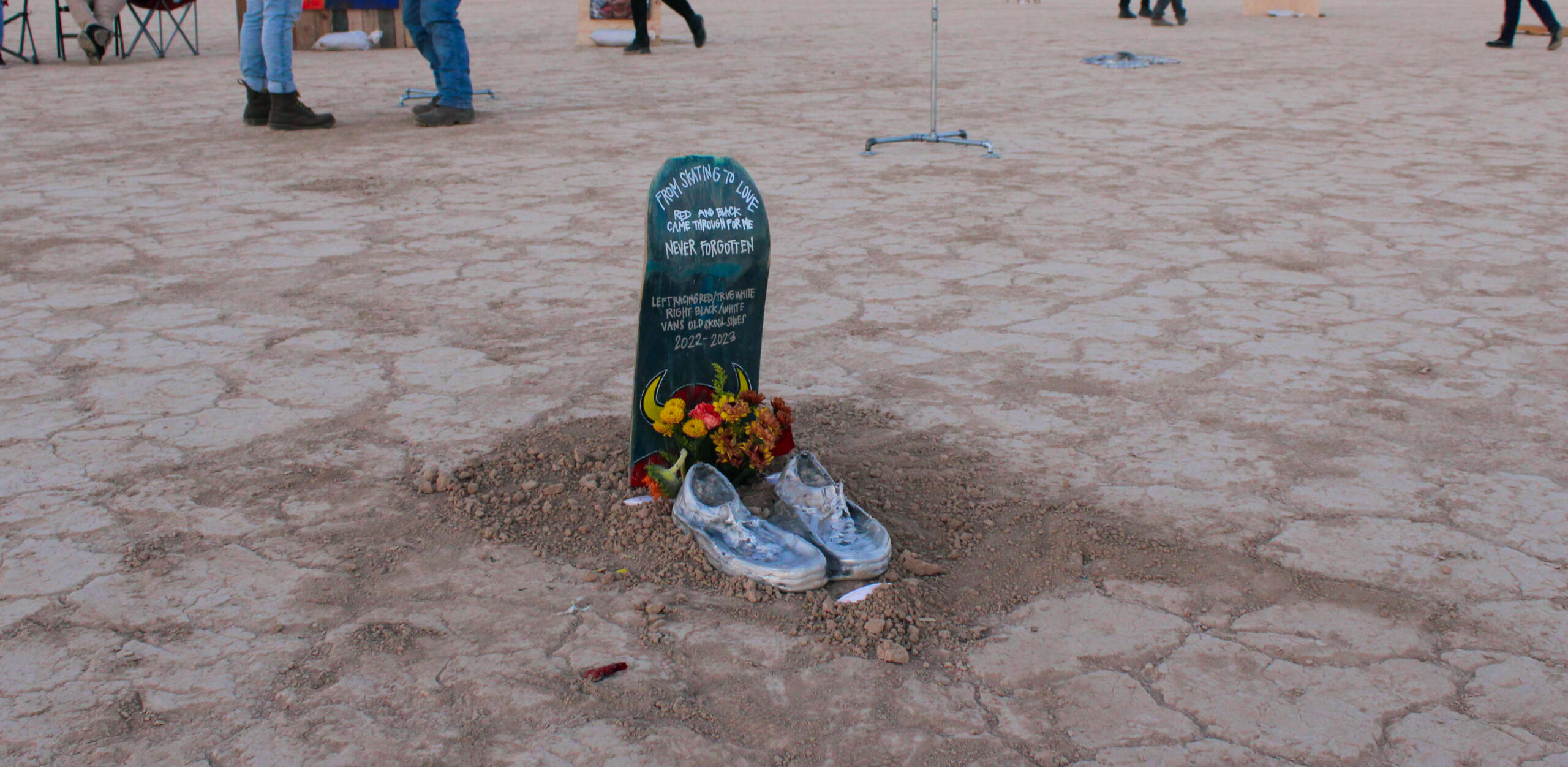 Image of sculpture by Dan45 Hernandez. Features skateboard attached vertically in ground in the shape of a tombstone. At the base of the "tombstone", there are flowers and artist's shoes cast in aluminum. Skateboard includes memorial inscription.