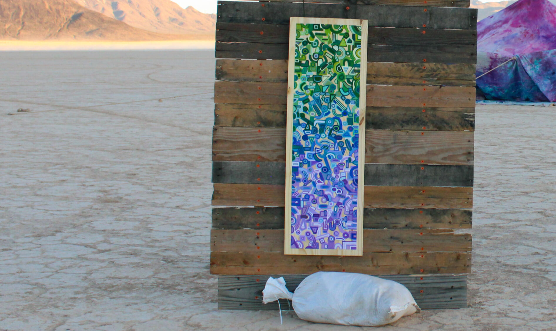 Image of painting by Maitlyn Holloway. Features painted abstract shapes in vertical gradient from green to purple. Painting is on a wooden board and hung on a temporary wall.