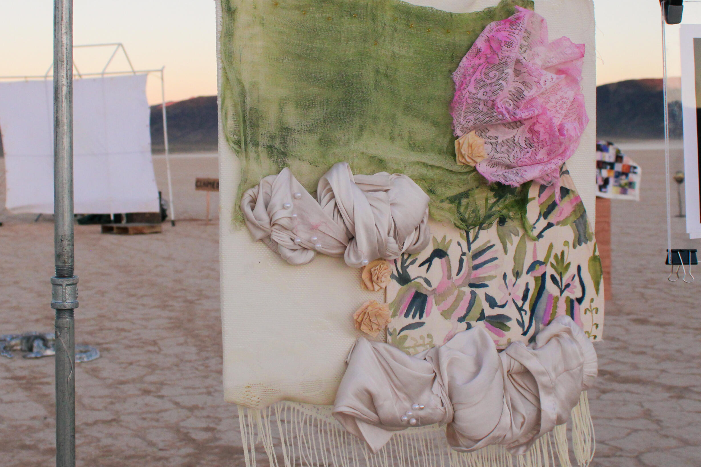 Image of sculpture by Haide Callejas. Features Otomi artisan embroidery on a suspended reclaimed fabric banner. The fabric is dyed pink and green and assembled in a garden-scape visage. Piece is supended from a clothesline.