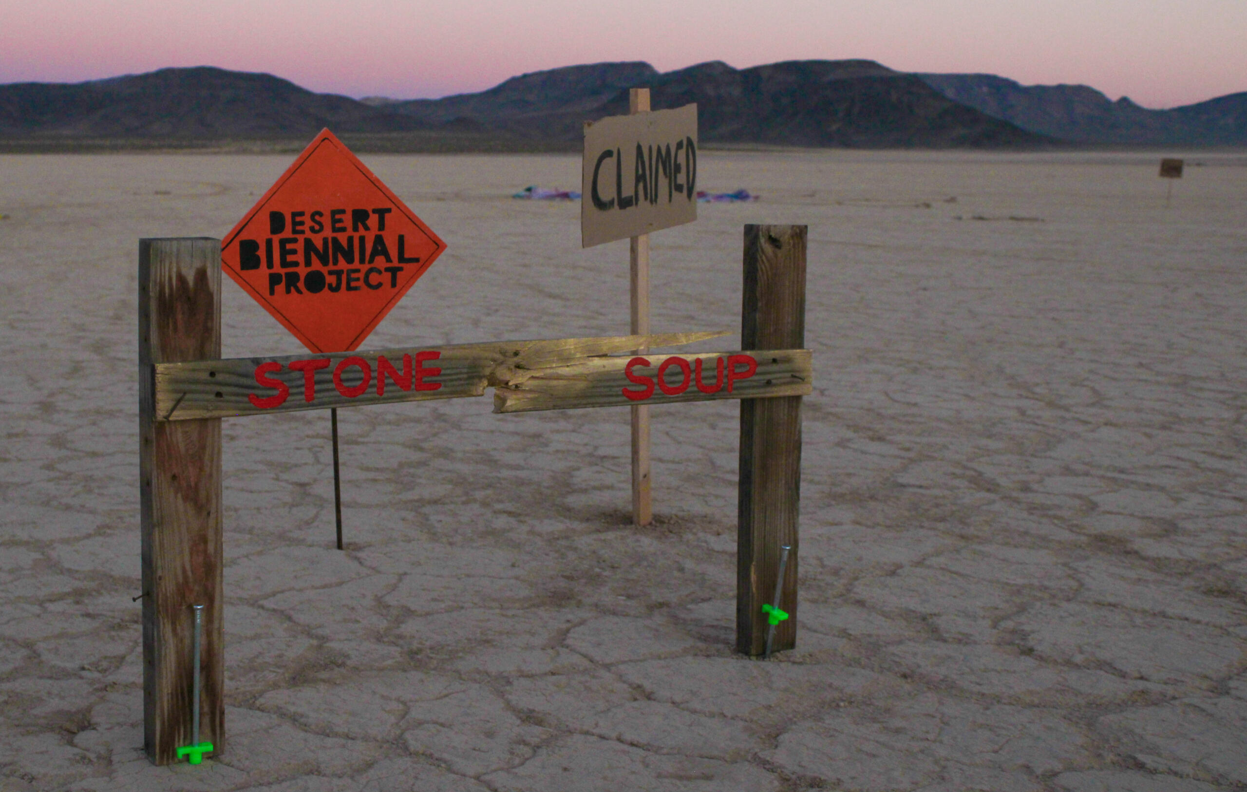 Image from Stone Soup event from 2023 Desert Biennial Project. Features bright orange diamond sign with black lettering spelling out Desert Biennial Project behind a sign made from dimensional lumber with Stone Soup painted in red on front of sign.