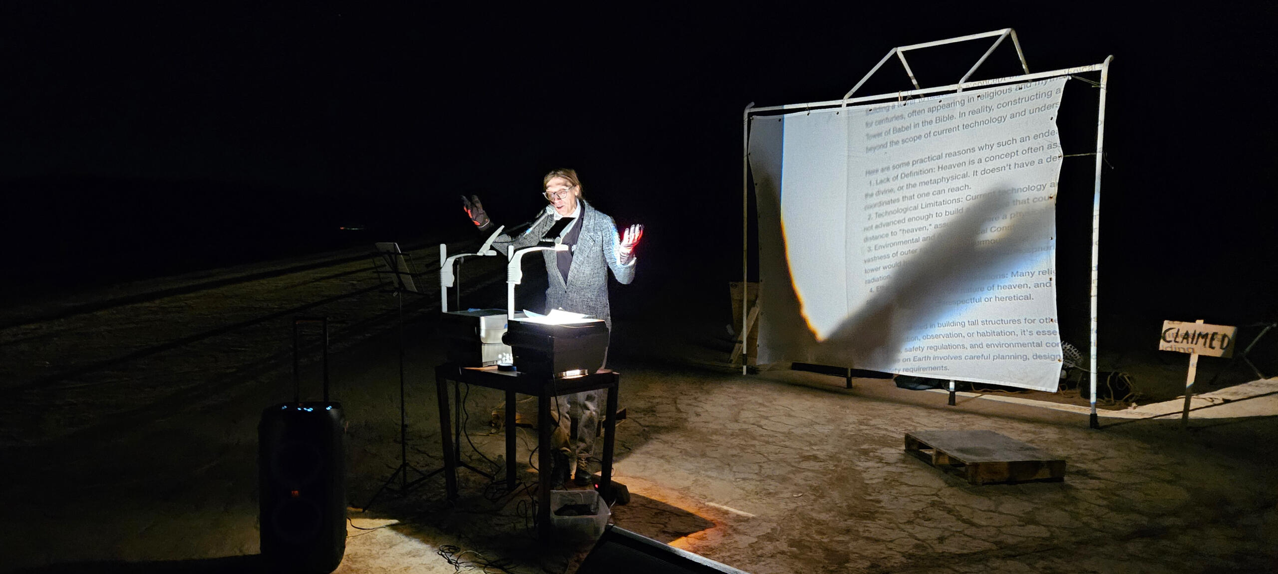 Still from performance by Keeva Lough. Features Keeva standing with arms facing up bent at elbows in front of two projectors that are projecting excerpts of conversation with ChatGPT about the Tower of Babel onto white stage behind her.