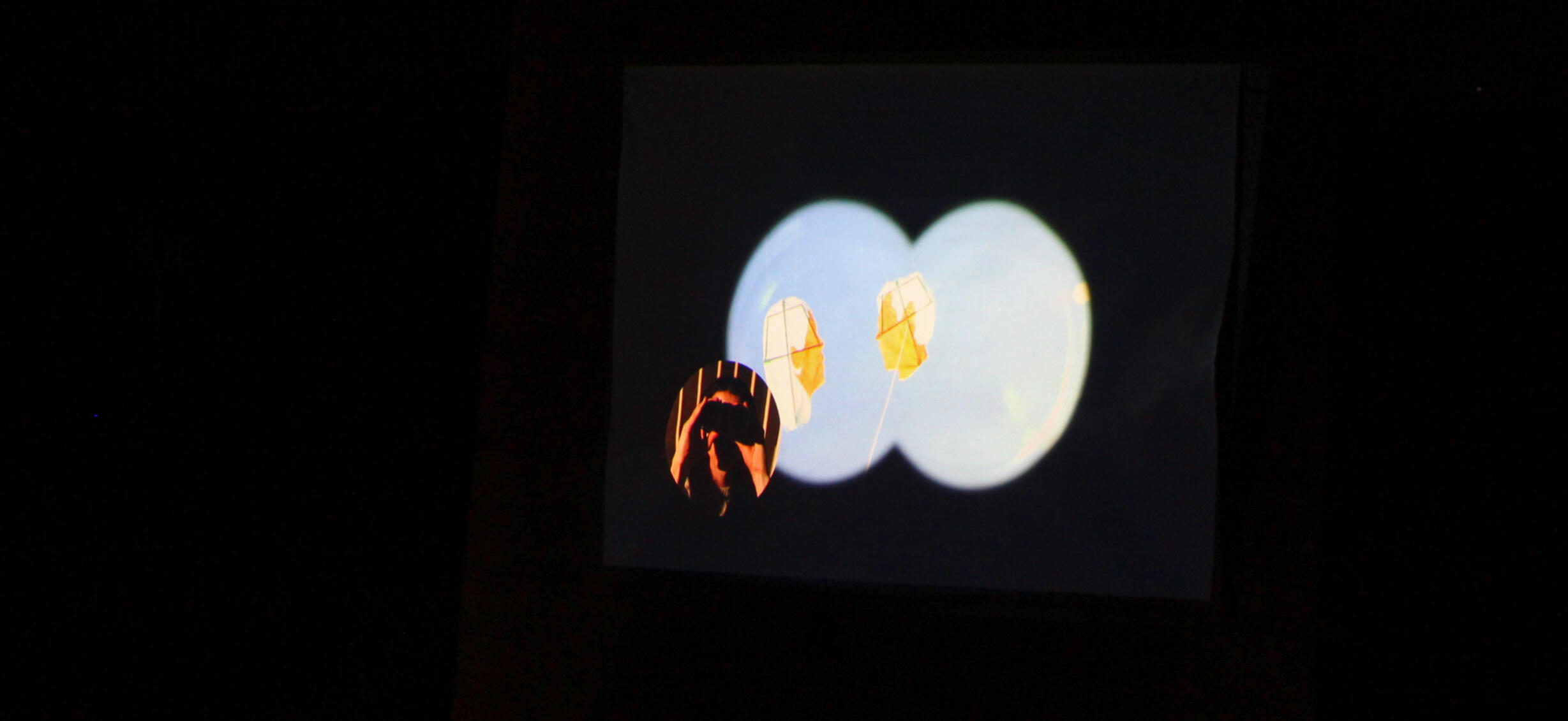 Still from video by Lane Sheehy. Features binocular view for two kites floating in the sky with a smaller circle to the left of the artist holding binoculars to her eyes. Piece is photographed at night and projected onto a temporary wall.