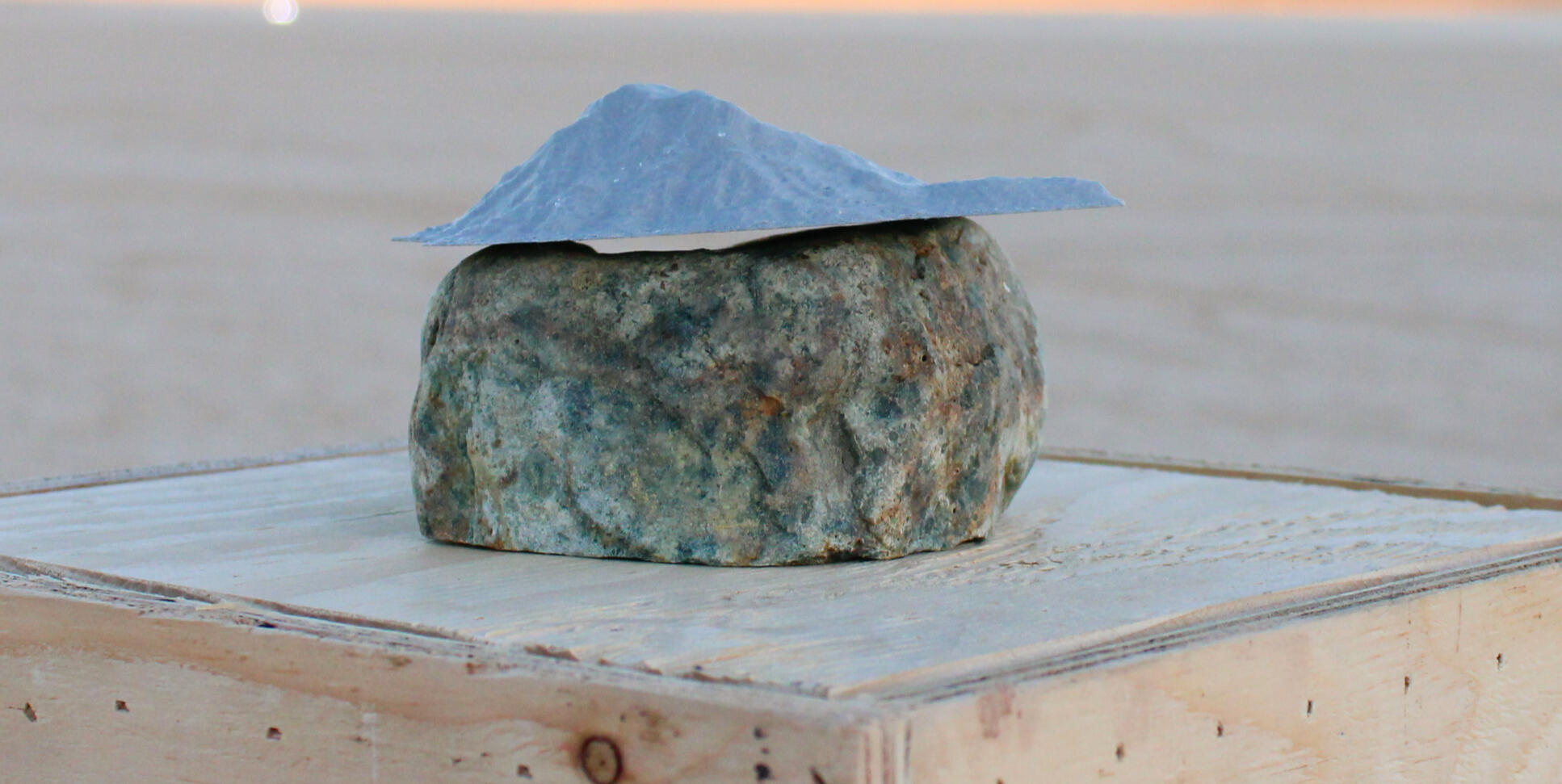 Image of sculpture by Luke Rizzotto. Features rock with 3D-printed rock with flat base sitting on top of it. Piece is displayed on top of wooden pedestal.