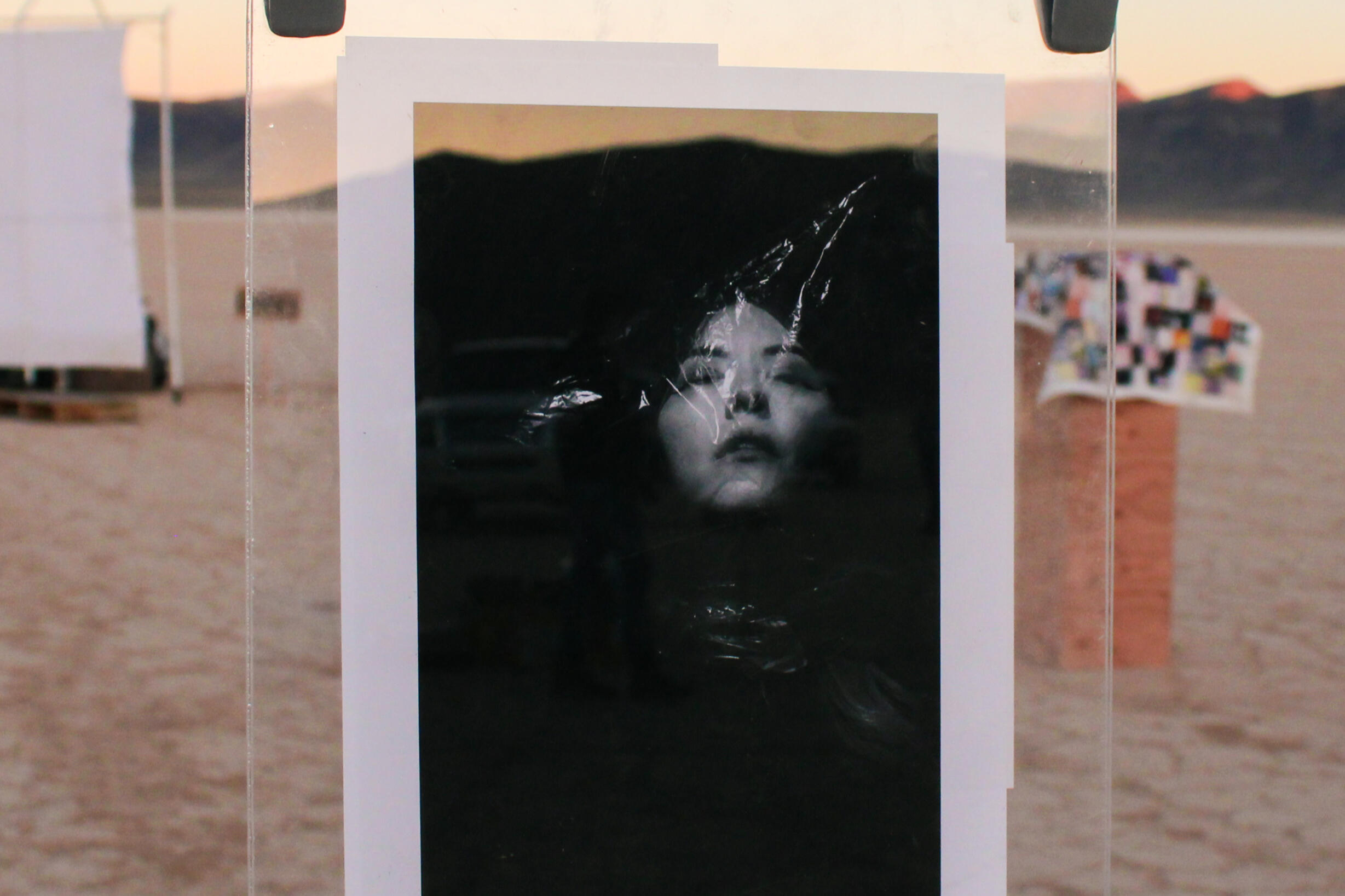 Image of photo by McKenzie Easter. Features black and white self-portrait of artist with clear plastic bag on head. Print is suspended between glass on clothesline.