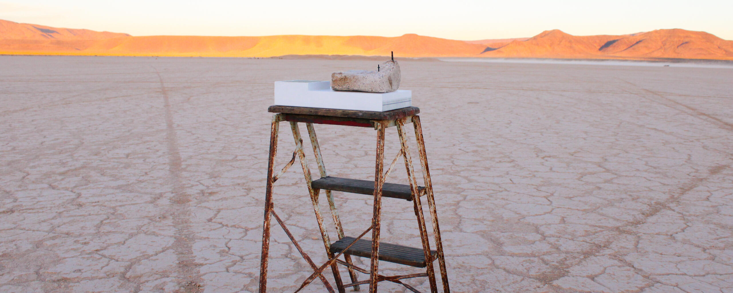 Image of sculpture by Mary Sabo. Features rock on white pedestal atop a stepladder with miniature ladder on top of rock and miniature cross slightly lower. Alongside rock is a pedestal with an illustrated shape and a cartoon glove hand pointing up.