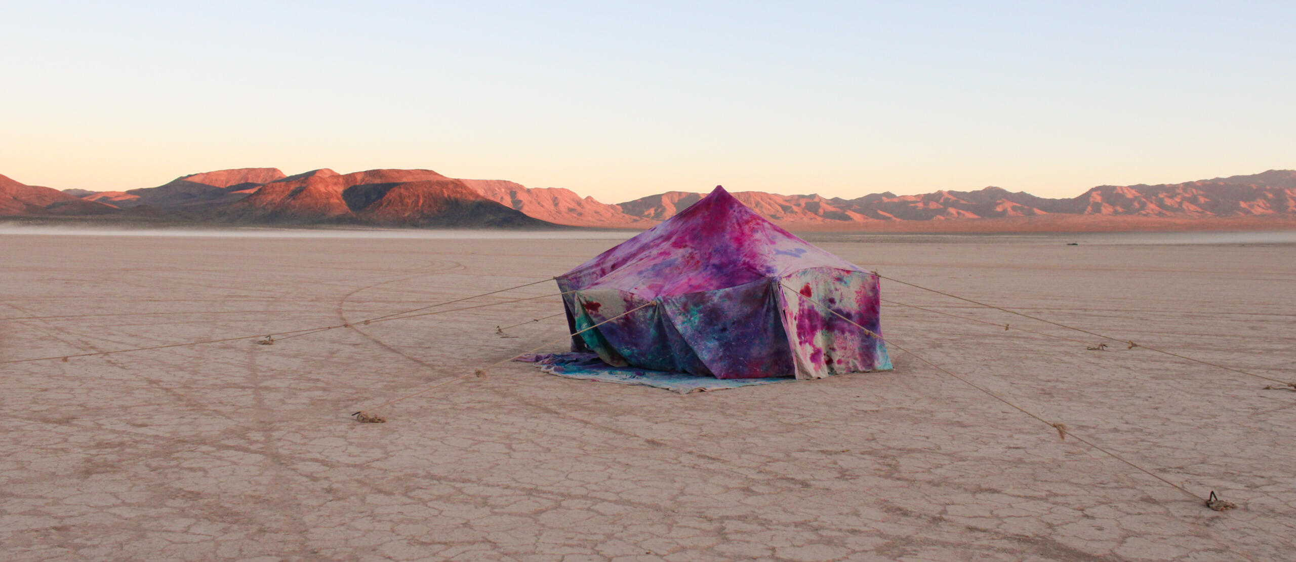 Image of sculpture by Karla Lagunas. Features tent stained with pink/purple/red/blue dye, pitched in the middle and tied down in circular motion around edges, like a circus tent.